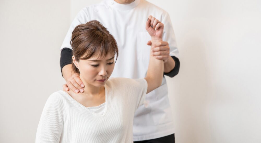 座った状態の女性の腕を持ち、【肩甲骨】周りや【肩関節】の動きを引き出している状況です。少し真剣な表情で自身の体と向き合っており、根深い肩こりや関節の可動域制限を検査しているようにも見えます。