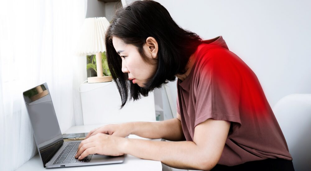 デスクワーク中に首を前に突き出した女性の背中が赤く発光している画像で、【頸部・背部(僧帽筋・肩甲挙筋・板状筋周辺)】における長時間の不良姿勢(ストレートネックや猫背)が引き起こす、ガンコな首こりや背中の強い張り、痛みの原因である猫背姿勢、ストレートネックを解説している様子。