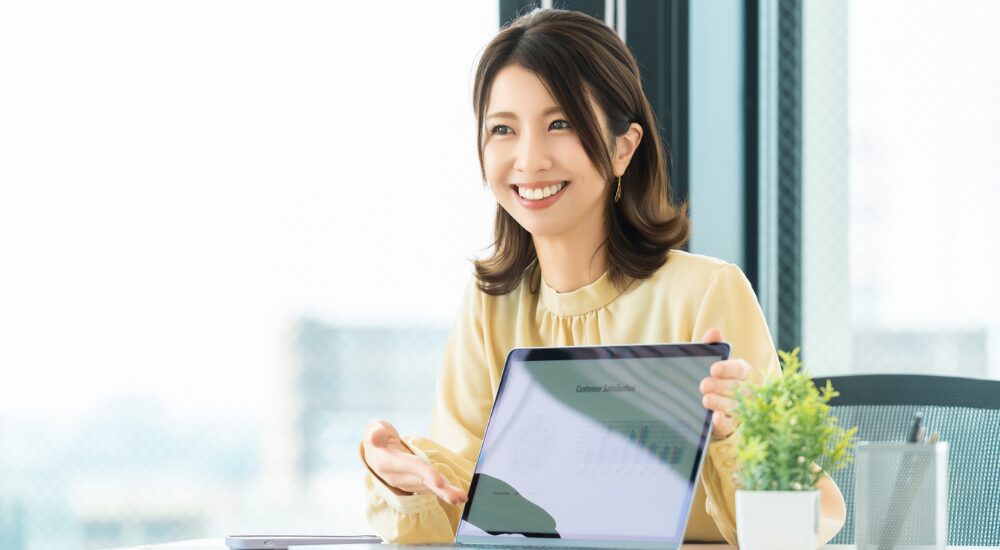 オフィスで女性が笑顔でノートパソコンのグラフ画面を見せながらプレゼンをしている画像で、【脳・全身（中枢神経・姿勢維持筋周辺）】を示し、首肩こりなどの不調が解消されたことで集中力がアップし、明るい表情で仕事のパフォーマンスが高まっている様子。